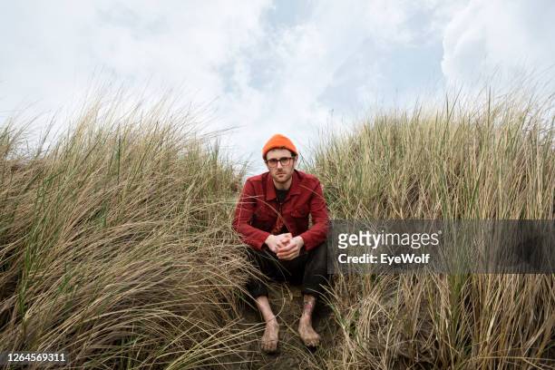 a young man sitting in the dunes with his hands crossed looking into camera. - wide stock-fotos und bilder