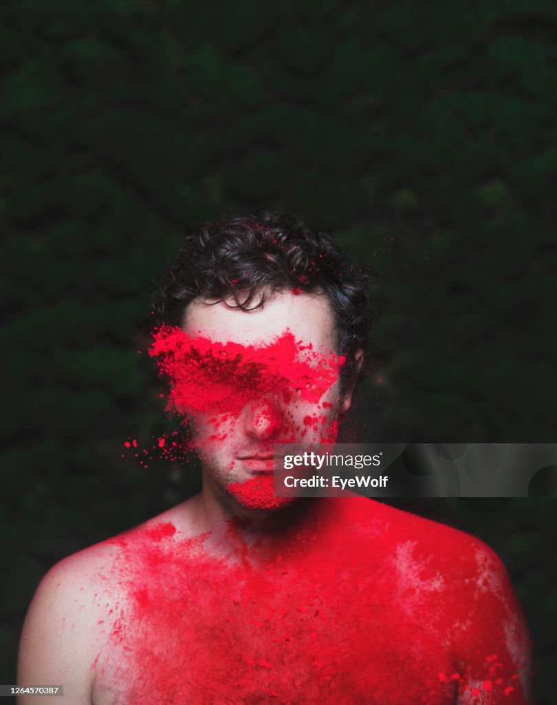 Man against a green background looking towards camera with Holi powder being thrown at him.