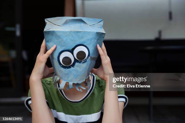 teenager with head in hands whilst wearing a blue painted paper bag and googly eyes over face - headache stock-fotos und bilder