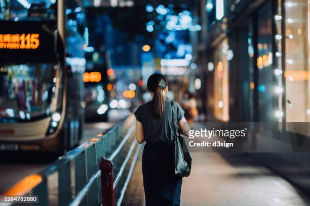 rear view of young asian woman walking on busy downtown city street after work at night, with busy traffic and illuminated city scene in background - evening walk stock-fotos und bilder
