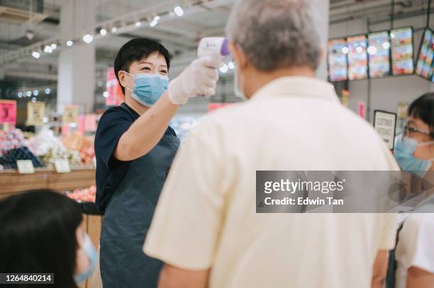 an asian chinese mid adult woman shopkeeper measuring senior man forehead taking temperature before entering supermarket as new standard operating procedure for covid 19 safety precaution - biosecurity stock pictures, royalty-free photos & images