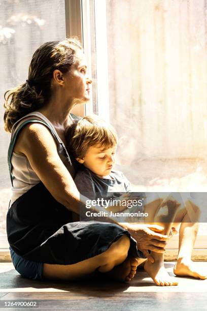 mature woman posing with her son, sitting on the floor, very sad looking through window worried about loss of her job due covid-19 pandemic - family sadness stock pictures, royalty-free photos & images