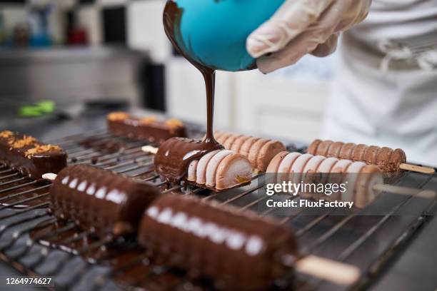 chef de postres cubriendo una galleta de pastel en una paleta con un chocolate negro derretido en una bandeja de red de metal - fábrica de chocolate fotografías e imágenes de stock