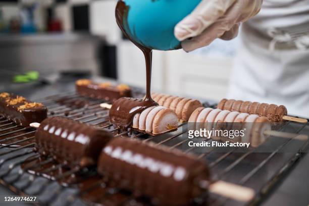 dessertchef die een cakekoekje op een popsickle met een gesmolten zwarte chocolade op een metaal nettodienblad behandelt - candy factory stockfoto's en -beelden