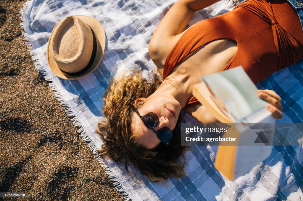 Mulher deita na praia lendo um livro