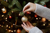 Woman decorating christmas tree with shiny golden bauble closeup. Preparation for christmas time. Modern glitter ornament in hands on background of festive tree in lights. Happy holidays