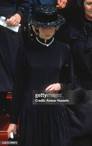 Diana, Princess of Wales, wearing a black dress and black boater hat with a netted veil and a heart shaped diamond necklace which was a gift from...