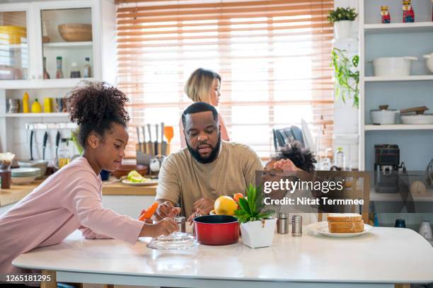 happy family sitting at a dining table with playing, talking together after breakfast while mother washing plate in background. - black mother and child cooking stock-fotos und bilder