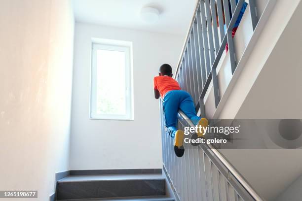 kleiner junge mit orangefarbenem sporthemd rutscht balustrade im treppenhaus hinunter - achtlos stock-fotos und bilder