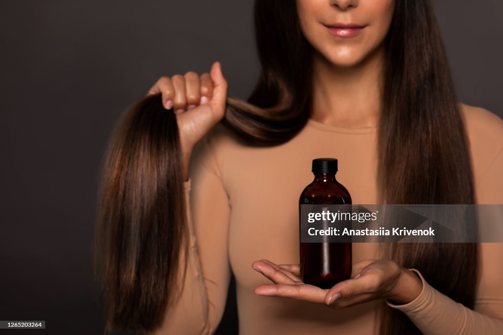 Brunette girl with long straight and shiny hair. Beauty skin woman holding her strong and healthy hair and oil or shampoo bottle over grey background. Cosmetic hair beauty salon concept.