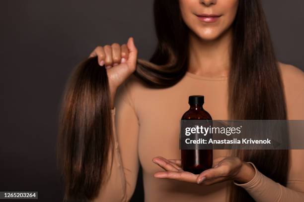 brunette girl with long straight and shiny hair. beauty skin woman holding her strong and healthy hair and oil or shampoo bottle over grey background. cosmetic hair beauty salon concept. - long hair - fotografias e filmes do acervo