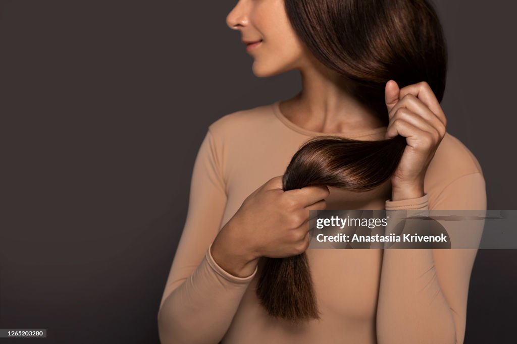 Brunette girl with long straight and shiny hair. Beauty skin woman holding her strong and healthy hair in her hands over grey background. Cosmetic hair beauty salon concept.