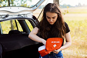 Young attractive girl stands near car with open back door and emergency lights is on, tries to find something in first aid kit