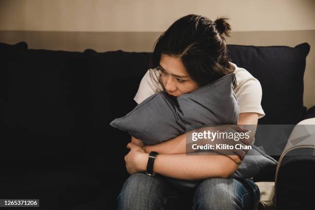 depressed asian woman sitting on sofa holding a cushion - besorgtheit stock-fotos und bilder