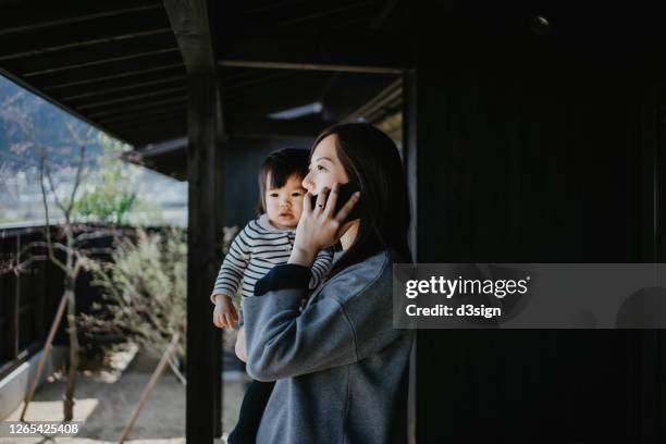 busy young asian mother talking on smartphone while taking care of cute little daughter carrying her on the balcony - リゾート　家族 ストックフォトと画像