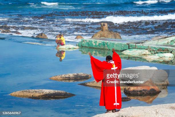 ein sangoma-heiler in einem roten gewand hebt die hände, um zu beten, bevor er die wassertaufe im atlantischen ozean, camp es bay, durchführt. - sangoma stock-fotos und bilder