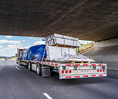 Red big rig semi truck transporting covered and tightened cargo on flat bed semi trailer running under the bridge across wide highway