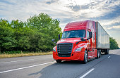 Red big rig long haul semi truck with black grille transporting cargo in dry van semi trailer running on the wide highway road