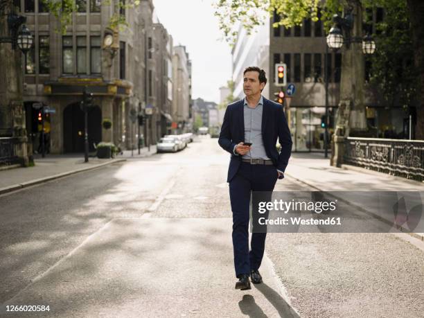 mature businessman walking on a city street holding smartphone - business men urban city stock-fotos und bilder