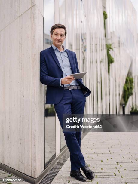 mature businessman with tablet leaning against a building in the city - anlehnen stock-fotos und bilder