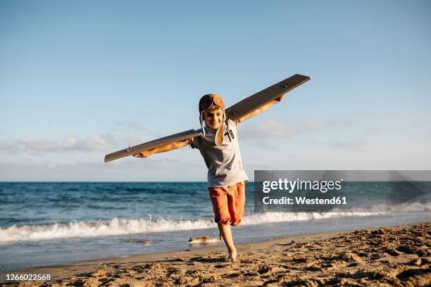 boy running with cardboard wings and aviator's cap at beach - 飛行機のまね ストックフォトと画像
