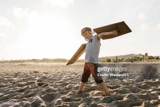 boy wearing aircraft wings and cap while standing with arms outstretched at beach - 飛行機のまね ストックフォトと画像
