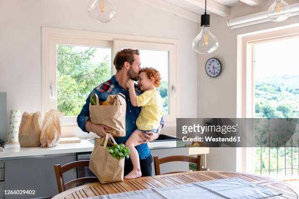 man kissing son while holding groceries bag in kitchen at home - young man groceries kitchen stock-fotos und bilder