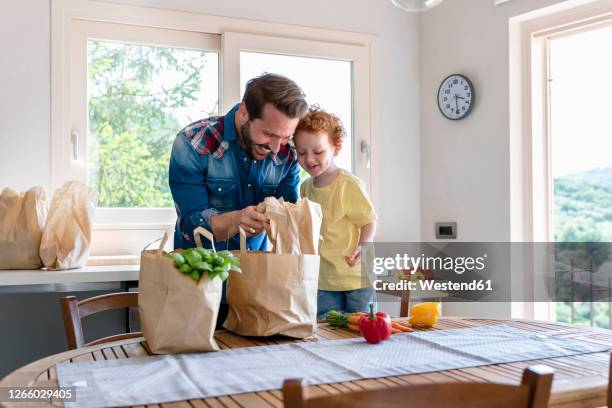smiling man showing groceries to son in kitchen at home - young man groceries kitchen stockfoto's en -beelden