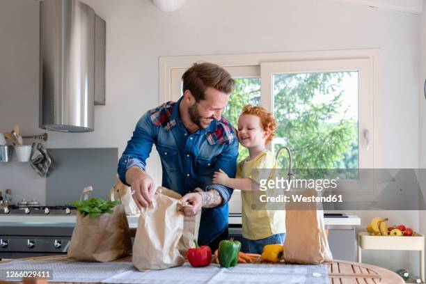 cheerful father and son with groceries standing at dining table in kitchen - young man groceries kitchen stockfoto's en -beelden