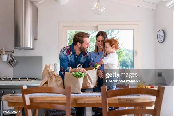 happy family with groceries bag at dining table in kitchen - tre quarti foto e immagini stock