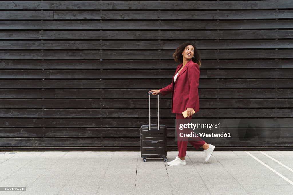 Young businesswoman looking back while walking with luggage on footpath by black wooden wall in city