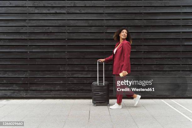 young businesswoman looking back while walking with luggage on footpath by black wooden wall in city - business woman looking over shoulder stock pictures, royalty-free photos & images