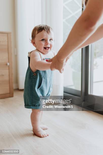 baby girl holding mother's hands while learning to walk on floor at home - einjährig stock-fotos und bilder