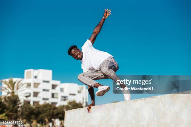 african man jumping over retaining wall against clear blue sky during sunny day - man jump outdoor young city stock pictures, royalty-free photos & images