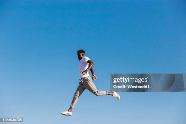 young man jumping against clear blue sky during sunny day - saltar actividad física fotografías e imágenes de stock