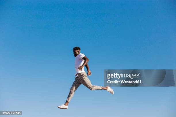young man jumping against clear blue sky during sunny day - saltare foto e immagini stock