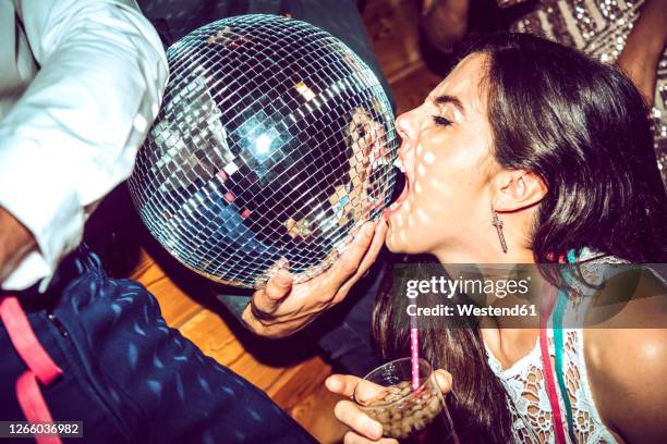 young woman with eyes closed holding drink and biting disco ball in glamorous party - diskokugel stock-fotos und bilder