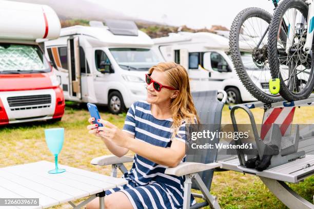 relaxed woman sitting next to camper enjoying vacation and using smartphone - campingstuhl stock-fotos und bilder