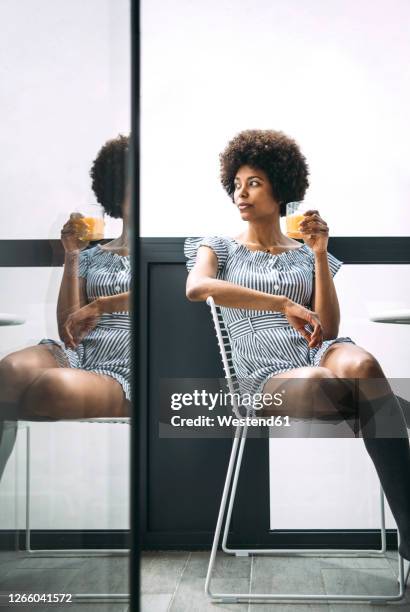thoughtful woman holding glass of orange juice while sitting on chair at balcony in penthouse - gestreiftes kleid stock-fotos und bilder