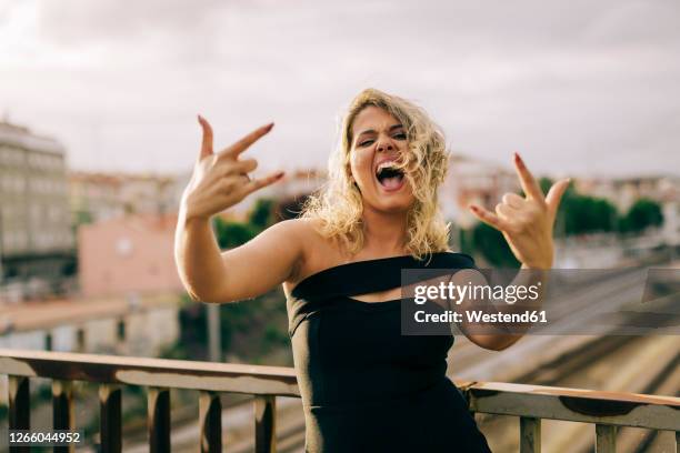 excited woman wearing black dress showing rock and roll sign in city - rock music ストックフォトと画像