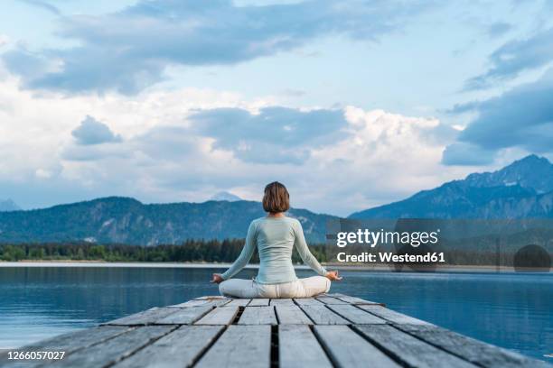 mid adult woman meditating while sitting on jetty over lake against cloudy sky - bergsteiger stockfoto's en -beelden