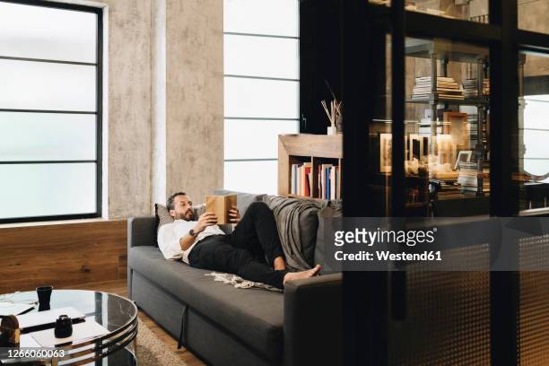 mature man lying on couch, relaxing, reading book - man living room fotografías e imágenes de stock