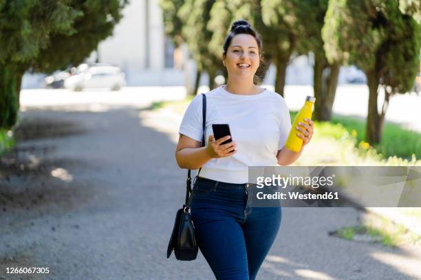 smiling curvy young woman with mobile phone and bottle in a public park - voluptuous woman stock-fotos und bilder