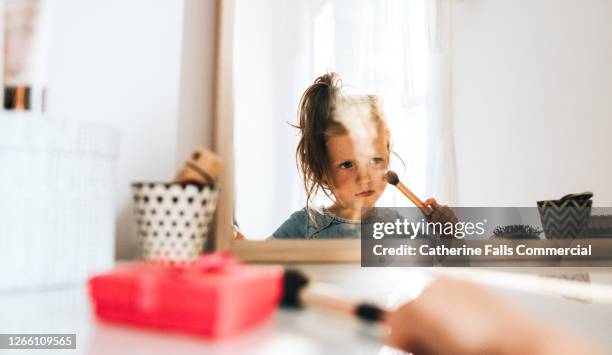 little girl in a bright white bedroom, looking at her reflection in a mirror, and playing with make-up. - adult imitation fotografías e imágenes de stock