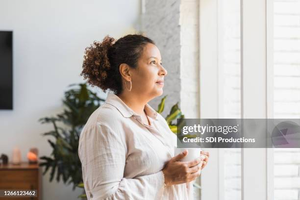 mid adult woman holds coffee while looking out window - woman praying stock pictures, royalty-free photos & images