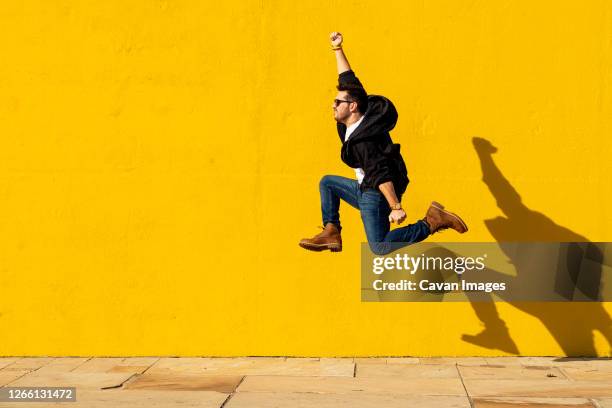 young man with sunglasses jumping in front of a yellow wall. - enthusiasm stock pictures, royalty-free photos & images