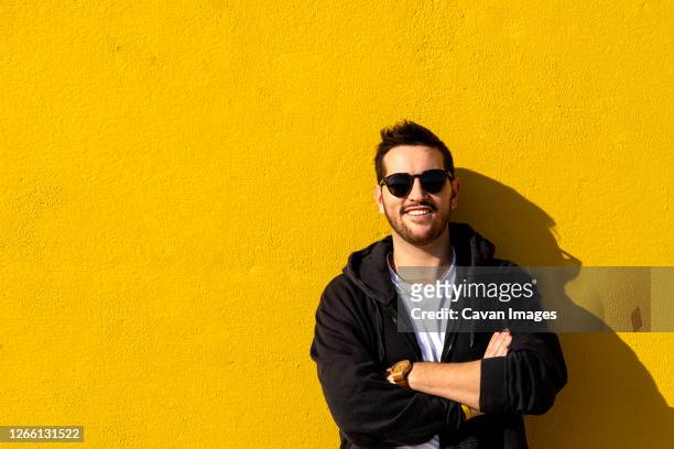 young bearded man standing against a yellow wall with crossed arms - geel jak stockfoto's en -beelden