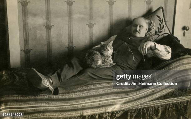 Rotund man laying on a bed holds a cat in his lap, circa 1895.