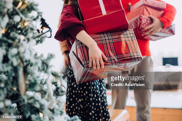 close up of young asian woman and man at the back holding a pile of wrapped christmas presents standing next to christmas tree preparing for a christmas party - red grant stock pictures, royalty-free photos & images