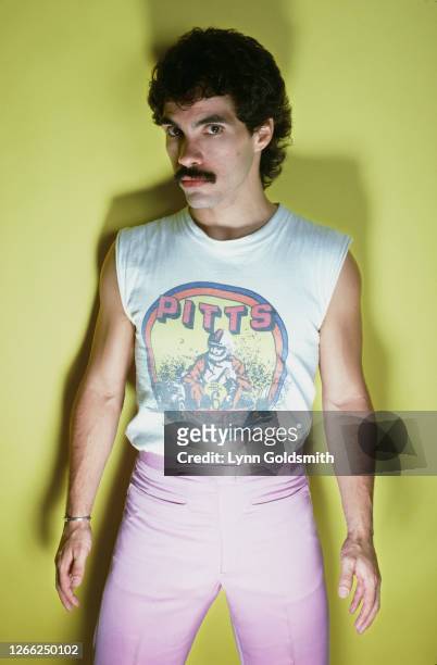 American singer-songwriter and guitarist John Oates wearing a white sleeveless Pitts go-kart t-shirt, with pink trousers, standing against a yellow...